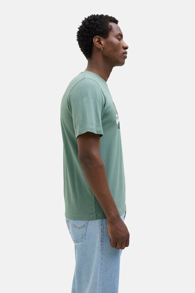 Kaki T-shirt met korte mouwen van JACK & JONES, gedragen met een lichtblauwe jeans en een gouden oorbel.