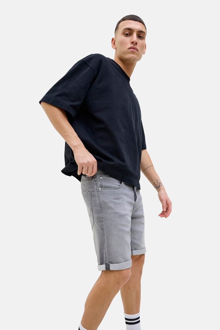 Korte spijkerbroek in grijs van JACK & JONES, gedragen met een oversized zwart T-shirt en gestreepte sokken.