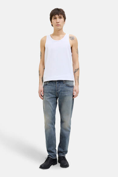 Taps toelopende jeans in middinblauw denim van JACK & JONES, gedragen met een witte tanktop en zwarte schoenen.
