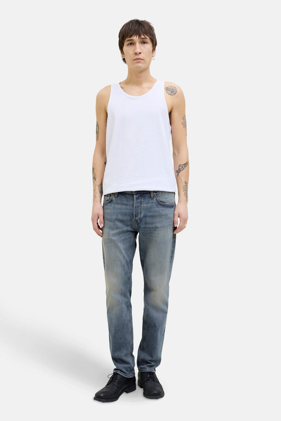 Taps toelopende jeans in middinblauw denim van JACK & JONES, gedragen met een witte tanktop en zwarte schoenen.
