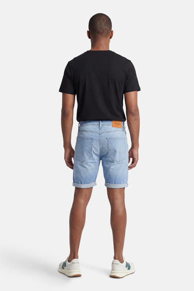 Short denim bleu clair de JACK & JONES, avec ourlets retroussés, porté de dos avec un t-shirt noir et des baskets crème à détails verts.