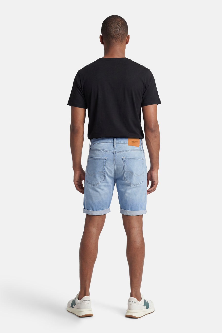 Short denim bleu clair de JACK & JONES, avec ourlets retroussés, porté de dos avec un t-shirt noir et des baskets crème à détails verts.