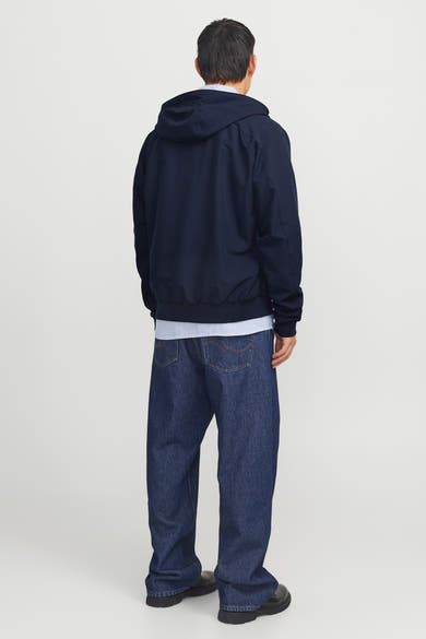 Donkerblauwe regenjas van JACK & JONES, achterkant, met capuchon en gedragen over een gestreept overhemd en een blauwe spijkerbroek.