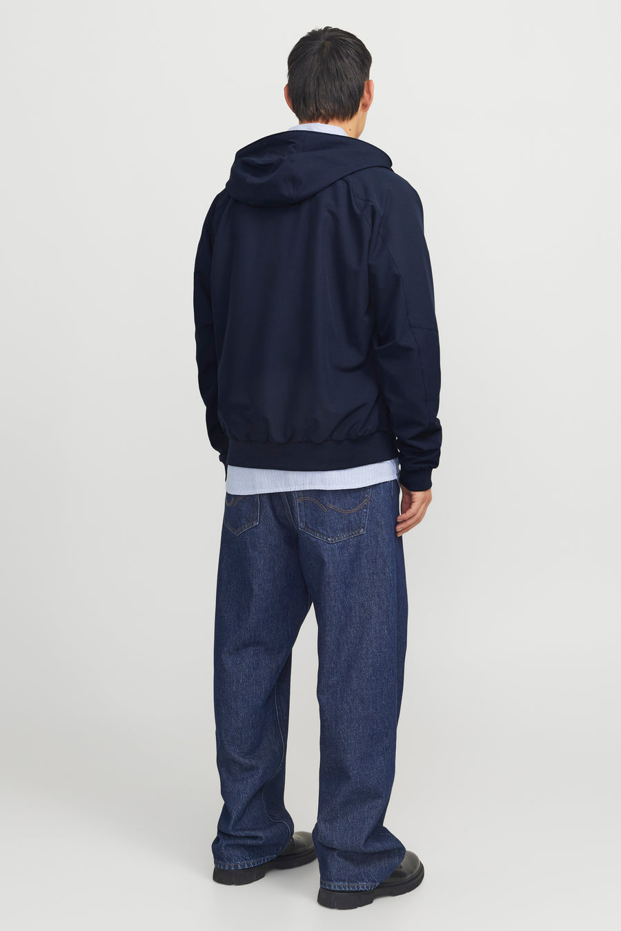 Donkerblauwe regenjas van JACK & JONES, achterkant, met capuchon en gedragen over een gestreept overhemd en een blauwe spijkerbroek.