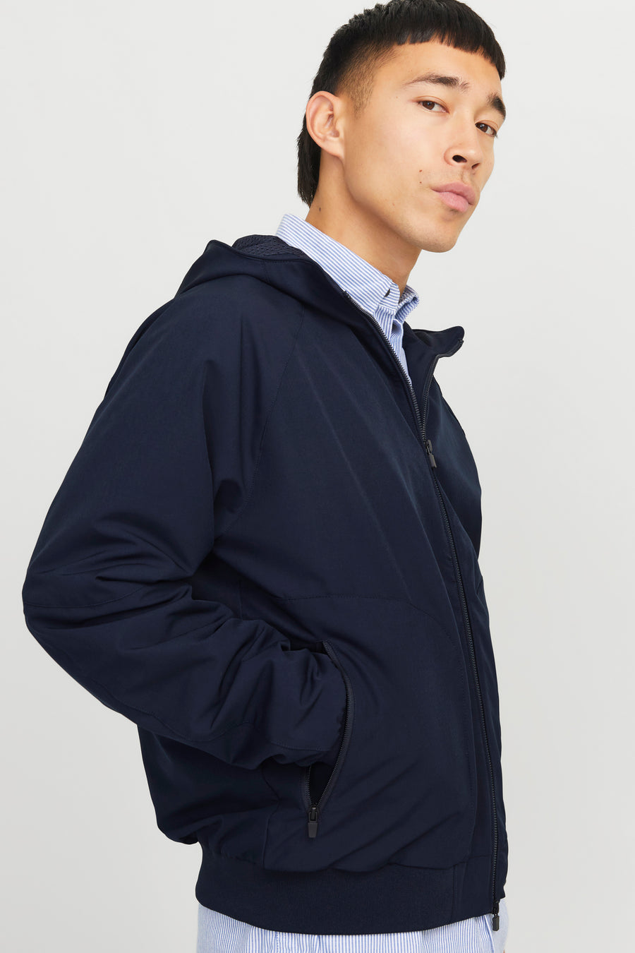 Donkerblauwe regenjas van JACK & JONES, met capuchon en ritssluiting, gedragen over een gestreept overhemd.