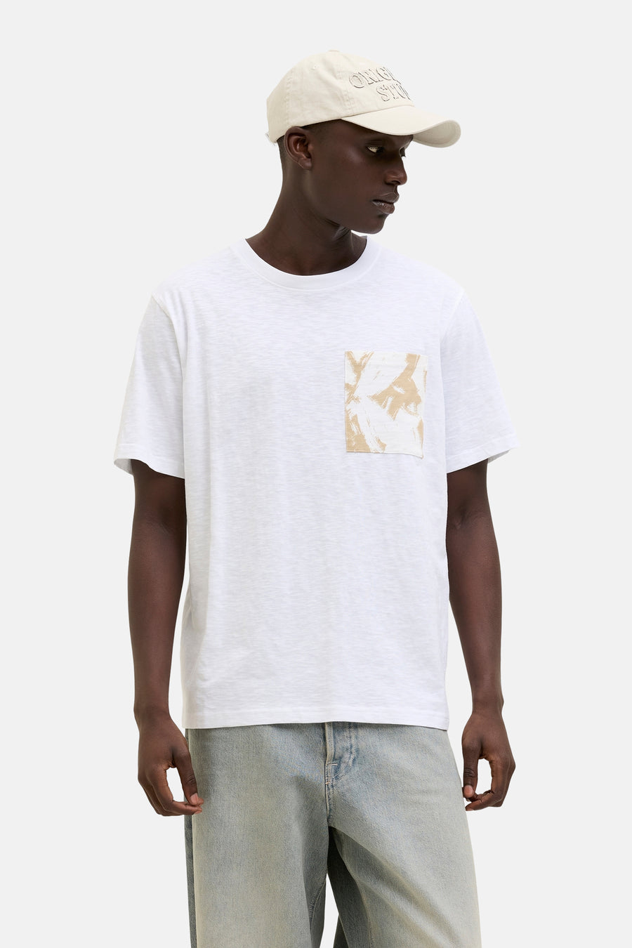 T-shirt blanc à manches courtes par ORIGINALS BY JACK & JONES avec poche imprimée abstraite, porté avec un jean et une casquette.
