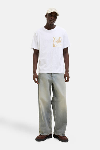 Wit T-shirt met korte mouwen van ORIGINALS BY JACK & JONES met abstracte opgedrukte zak, gedragen met een spijkerbroek en een pet.