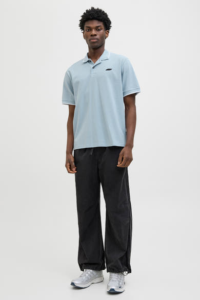 Polo bleu à manches courtes de JACK & JONES, porté avec un pantalon noir ample et des baskets argentées.