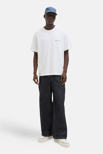 T-shirt blanc à manches courtes par ORIGINALS BY JACK & JONES, porté avec un pantalon denim ample et une casquette.
