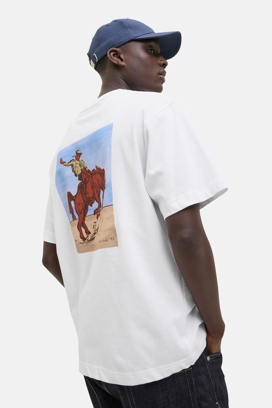 Wit T-shirt met korte mouwen van ORIGINALS BY JACK & JONES met een illustratie van een cowboy op de rug, gedragen met een blauwe pet en een spijkerbroek.