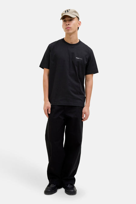 T-shirt noir à manches courtes par ORIGINALS BY JACK & JONES, porté avec un pantalon noir ample et une casquette beige.

