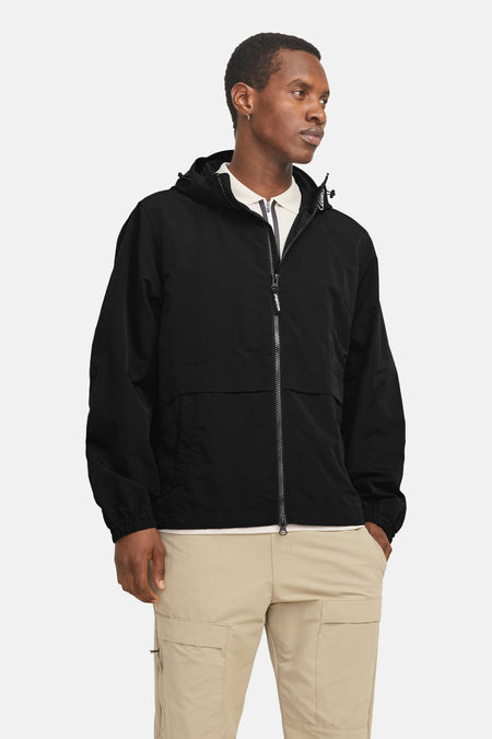 Kort zwart jack van JACK & JONES, met capuchon en volledige ritssluiting, gedragen over een poloshirt en een beige cargobroek.