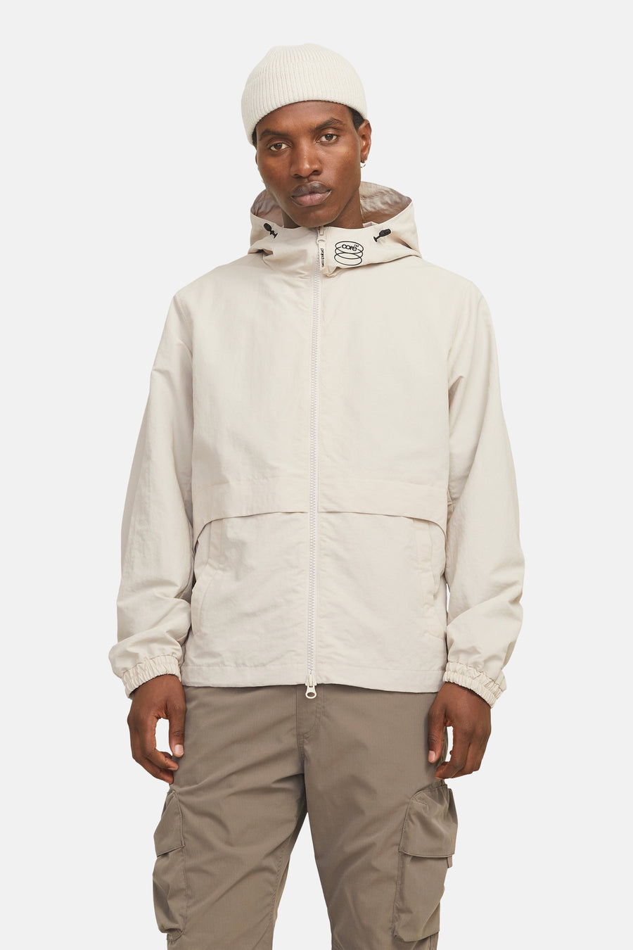 Korte beige jas van JACK & JONES, met capuchon en ritssluiting, gedragen met een crèmekleurige muts en een cargobroek.