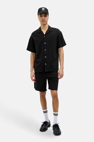 Chemise noire à manches courtes par ORIGINALS BY JACK & JONES, portée avec un short noir, des chaussettes rayées et un chapeau noir.