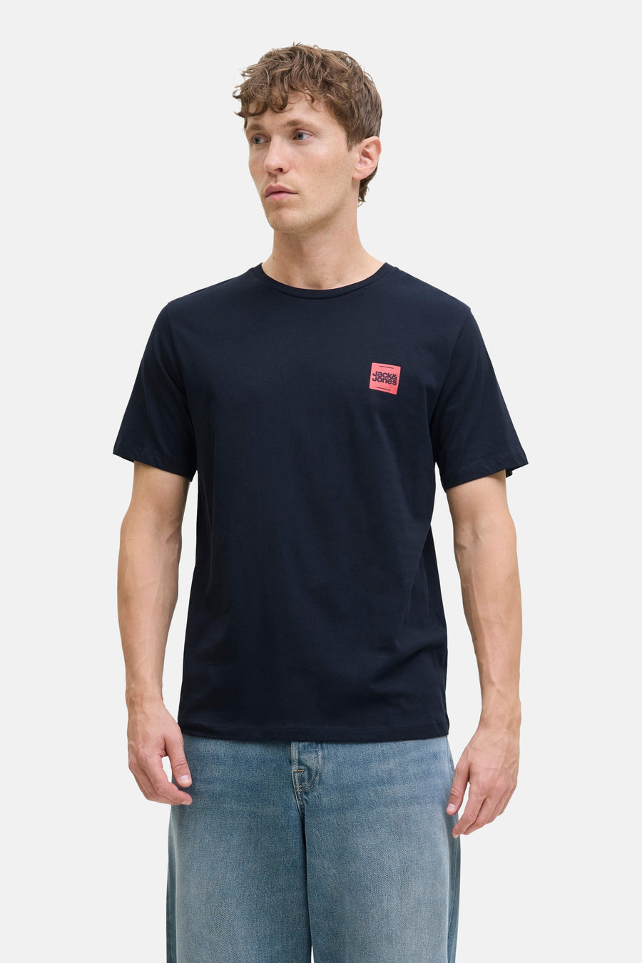 T-shirt bleu à manches courtes par ORIGINALS BY JACK & JONES, avec un logo carré rouge sur le devant, porté avec un jean.