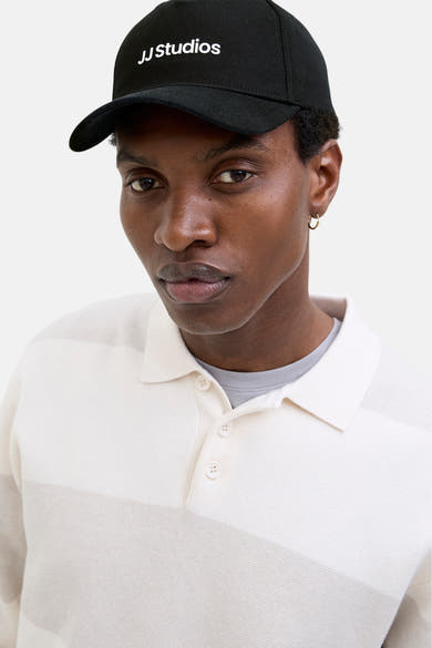 Casquette noire d’ACCESSORIES BY JACK & JONES, portée par un homme en polo rayé et boucles d'oreilles dorées.