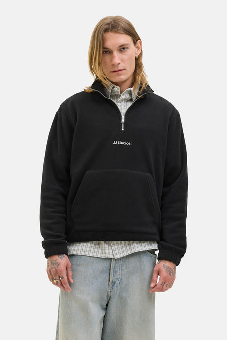 Zwarte fleece van JACK & JONES met een ritssluiting en een kangoeroezak op een man die een geruit overhemd en een spijkerbroek draagt.