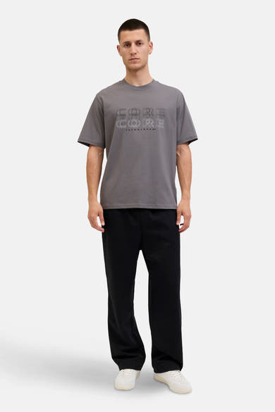 Grijs T-shirt met korte mouwen van JACK & JONES, met een grafische "CORE" print, gedragen met een zwarte broek en witte sneakers.