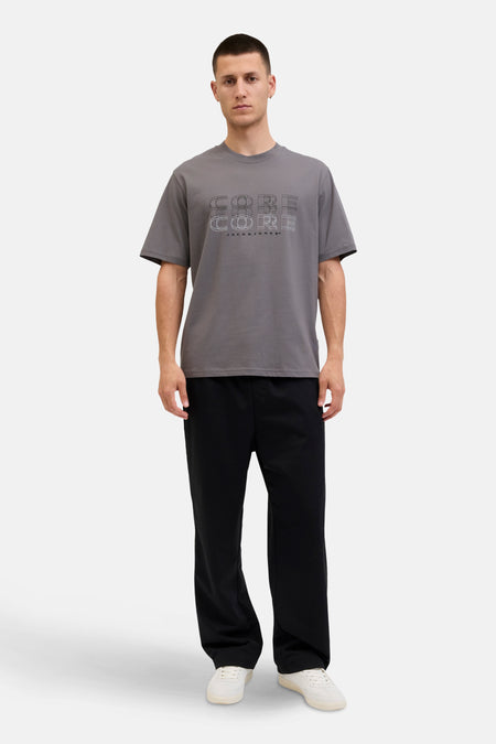 Grijs T-shirt met korte mouwen van JACK & JONES, met een grafische "CORE" print, gedragen met een zwarte broek en witte sneakers.