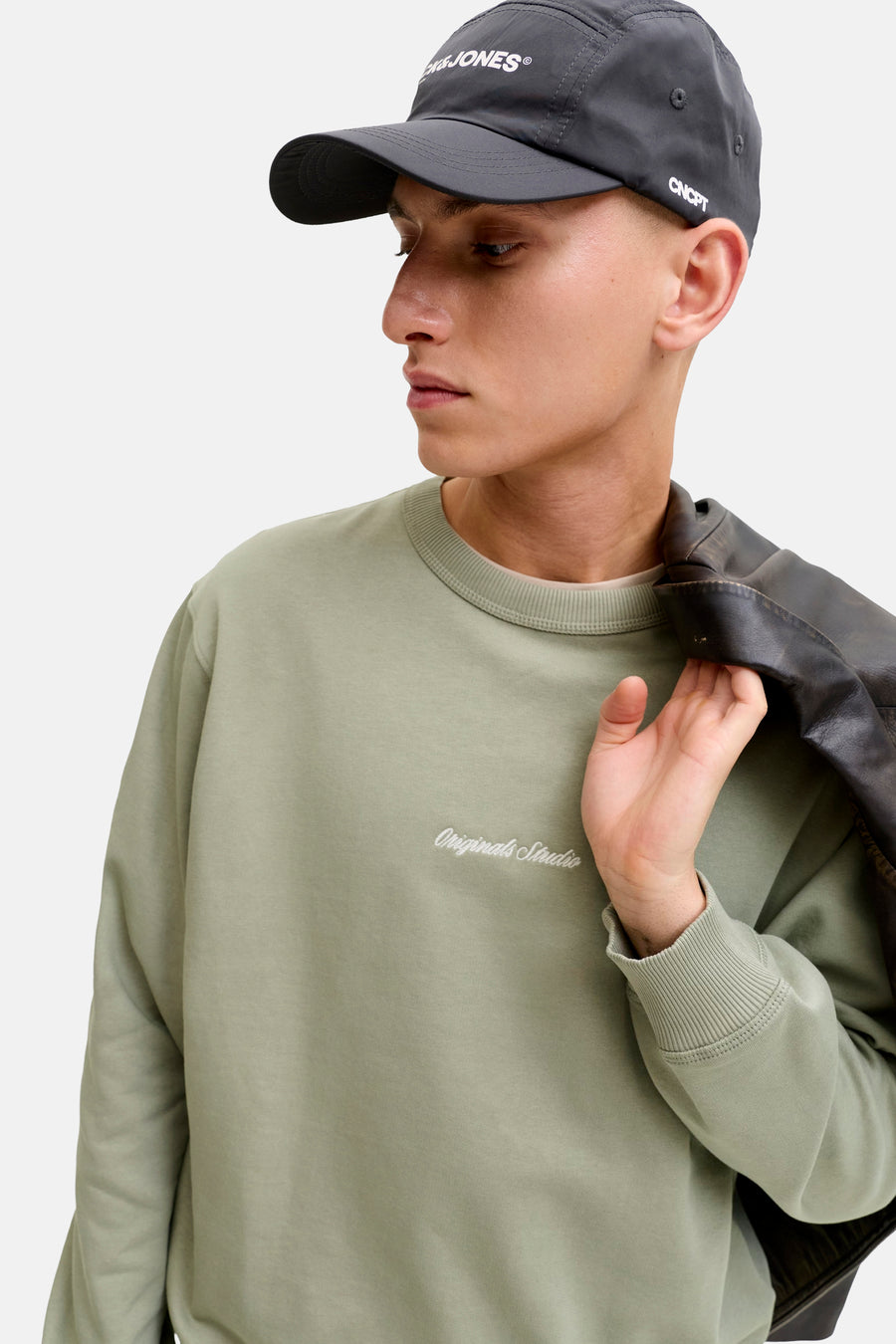 Groene rondehals sweatshirt van ORIGINALS BY JACK & JONES, gedragen met een leren jas over de schouder en een pet.