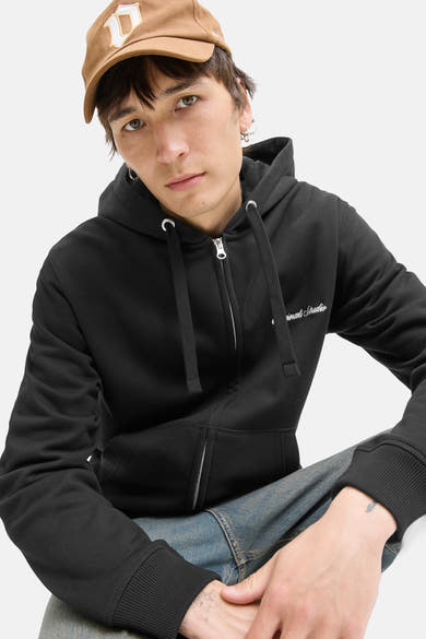 Zwarte rits-sweatshirt van PREMIUM by JACK & JONES, met capuchon en trekkoord, gedragen met een spijkerbroek en een bruine pet.