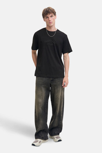 T-shirt noir à manches courtes de JACK & JONES, avec un motif ton sur ton sur le devant, porté avec un jean ample.
