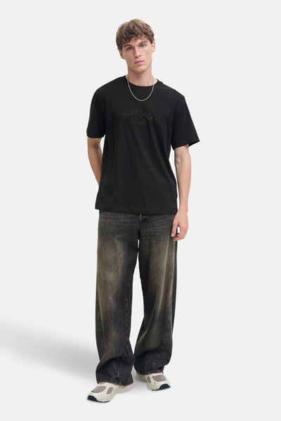 T-shirt noir à manches courtes de JACK & JONES, avec un motif ton sur ton sur le devant, porté avec un jean ample.
