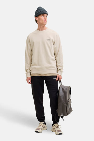 Sweat-shirt beige Originals by Jack & Jones, porté avec un pantalon de survêtement noir, un bonnet gris et un sac à dos en cuir.