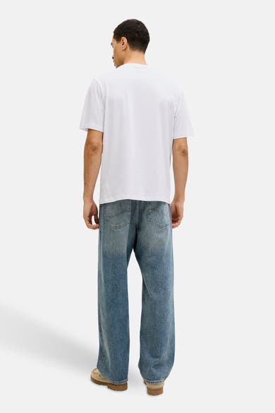 Wit T-shirt met korte mouwen van JACK & JONES, gedragen met een wijde, lichtblauwe spijkerbroek en lichtbruine schoenen.