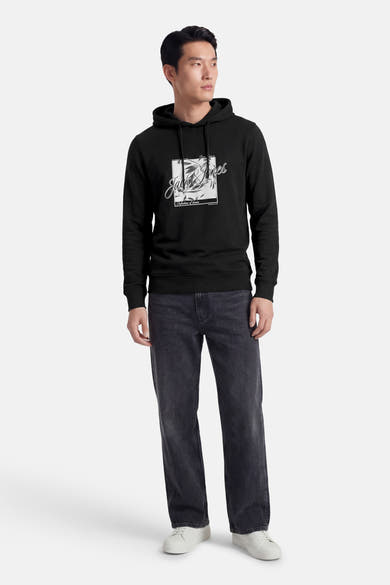 Zwarte hoodie van JACK & JONES, met een print op de voorkant, gedragen met een spijkerbroek en witte sneakers.