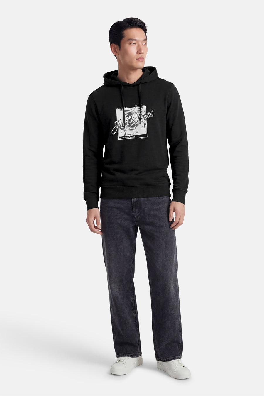 Zwarte hoodie van JACK & JONES, met een print op de voorkant, gedragen met een spijkerbroek en witte sneakers.