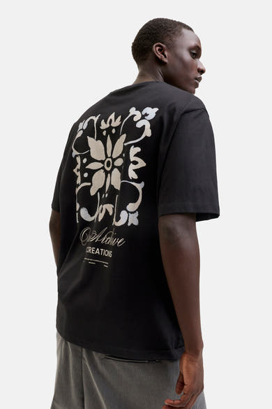 T-shirt noir de Originals by Jack & Jones avec grand motif floral abstrait blanc cassé imprimé au dos, porté par un homme en pantalon gris, vu de dos.