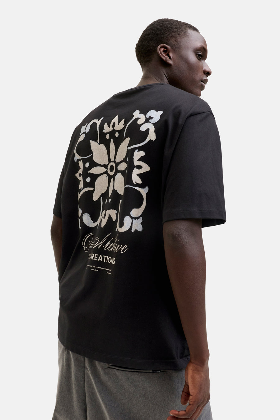 T-shirt noir de Originals by Jack & Jones avec grand motif floral abstrait blanc cassé imprimé au dos, porté par un homme en pantalon gris, vu de dos.