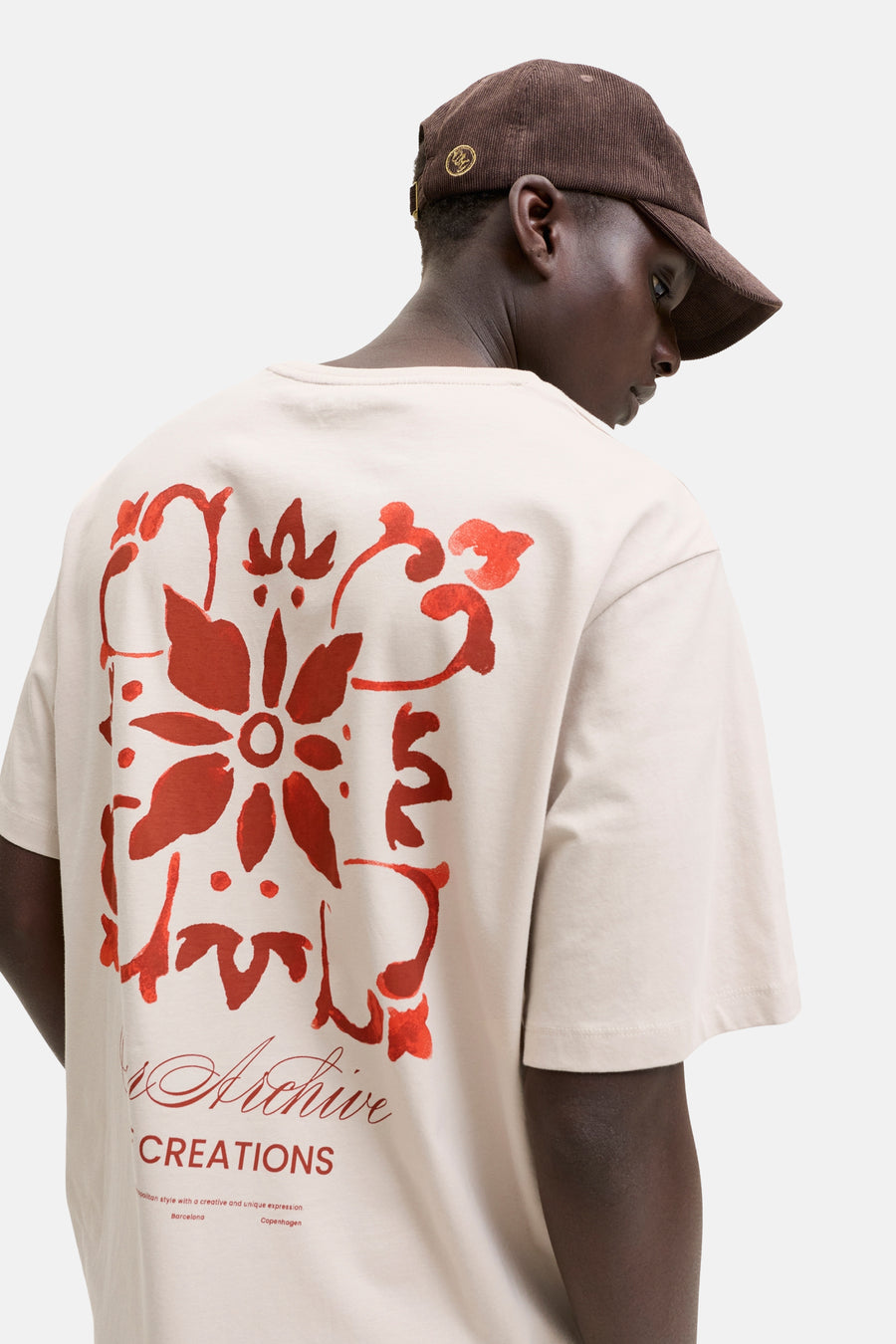 T-shirt beige de ORIGINALS BY JACK & JONES avec grand motif floral rouge et inscription "Archive CREATIONS" au dos. L'homme porte une casquette côtelée marron.