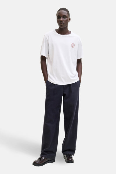 T-shirt blanc à manches courtes par ORIGINALS BY JACK & JONES, avec un logo rond sur la poitrine, porté avec un pantalon bleu marine.