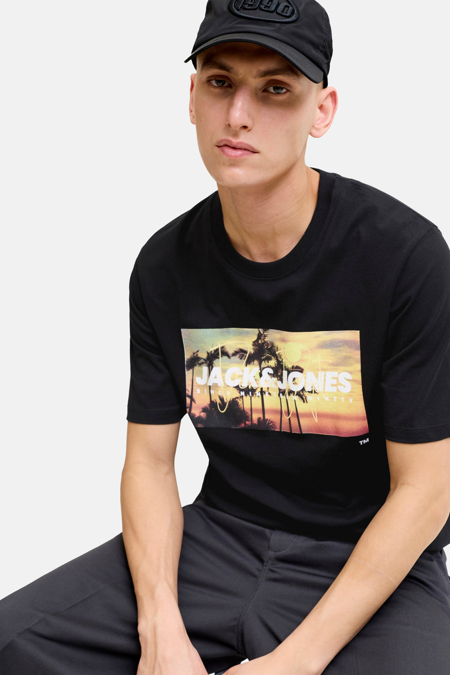 T-shirt noir de JACK & JONES avec imprimé graphique de palmiers au coucher de soleil, porté par un homme avec un pantalon foncé et une casquette noire.