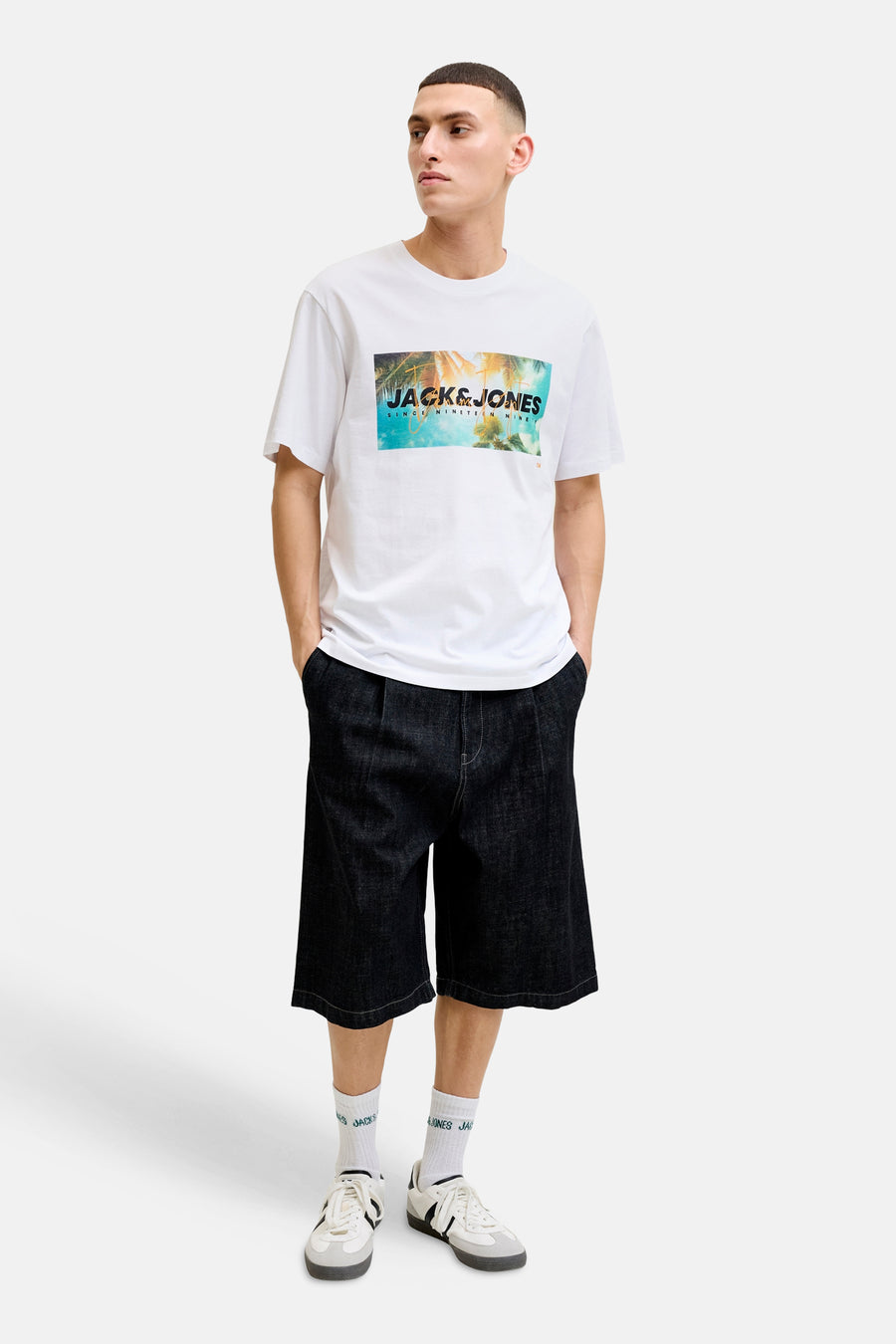 T-shirt blanc de JACK & JONES avec imprimé graphique de palmiers et logo, porté avec un short en jean.