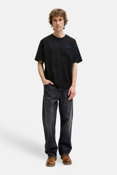 Black short-sleeve t-shirt with small blue chest embroidery, worn by model in full-length lifestyle view with dark jeans and brown boots.