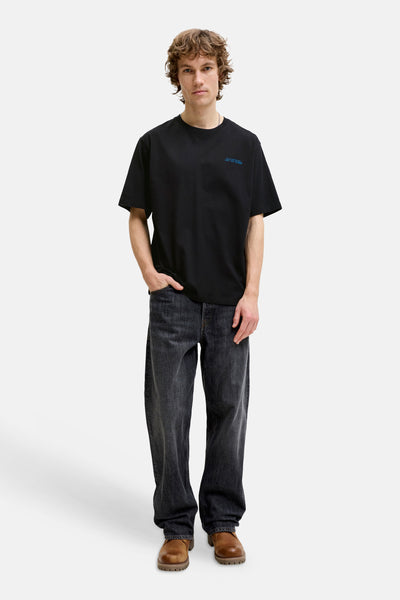 Black short-sleeve t-shirt with small blue chest embroidery, worn by model in full-length lifestyle view with dark jeans and brown boots.