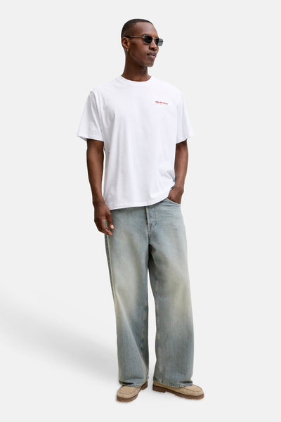 White short-sleeve t-shirt with small red logo on chest, worn by model in light blue jeans and tan loafers, front view