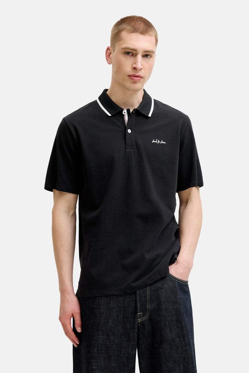 Zwart poloshirt van JACK & JONES met dubbele witte streep op de kraag, geborduurd Jack & Jones-logo en gedragen door een man in een spijkerbroek.