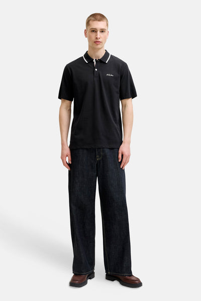 Zwart JACK & JONES poloshirt met witte bies op de kraag, witte knopen en geborduurd logo, gedragen met een wijde spijkerbroek en bruine loafers.