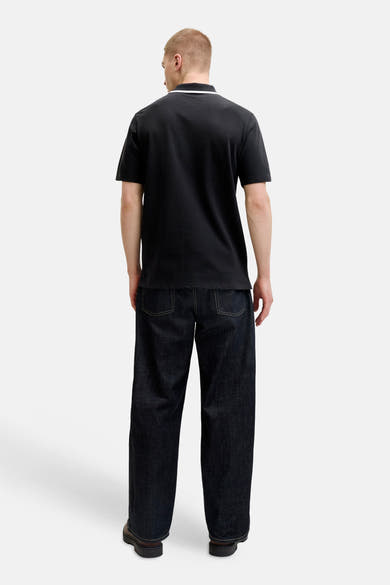 Polo noir de JACK & JONES avec col bordé de blanc, porté de dos avec un jean foncé à surpiqûres jaunes et des chaussures marron.