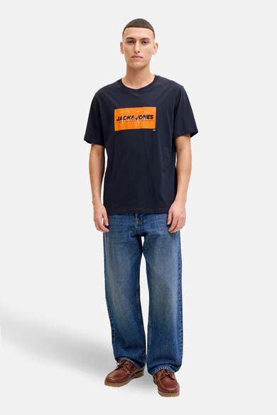 T-shirt bleu à manches courtes de JACK & JONES, avec un logo graphique orange sur le devant, porté avec un jean ample.
