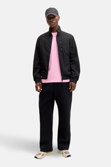 Zwarte regenjas van ORIGINALS BY JACK & JONES, gedragen met een roze T-shirt, een zwarte broek en beige sneakers.