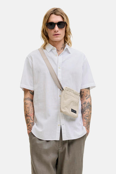 Chemise blanche à manches courtes de JACK & JONES, portée avec un sac bandoulière et un pantalon gris.
