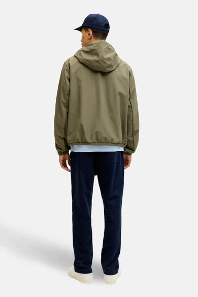 Korte groene jas van ORIGINALS BY JACK & JONES, met een capuchon, gedragen met een marineblauwe broek en witte sneakers.