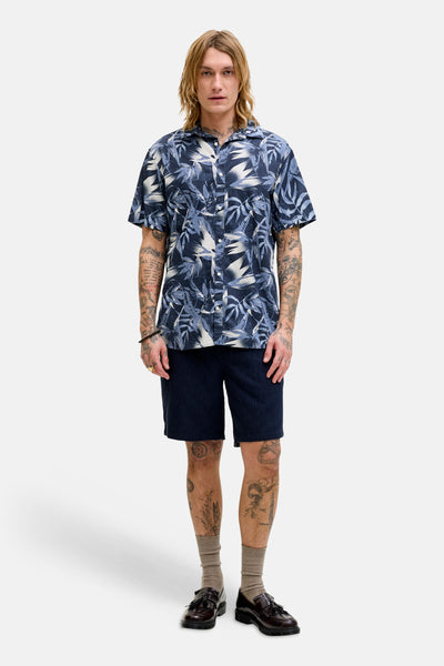 Chemise bleue à manches courtes de JACK & JONES, avec un motif de feuilles tropicales, portée avec un short et des mocassins.