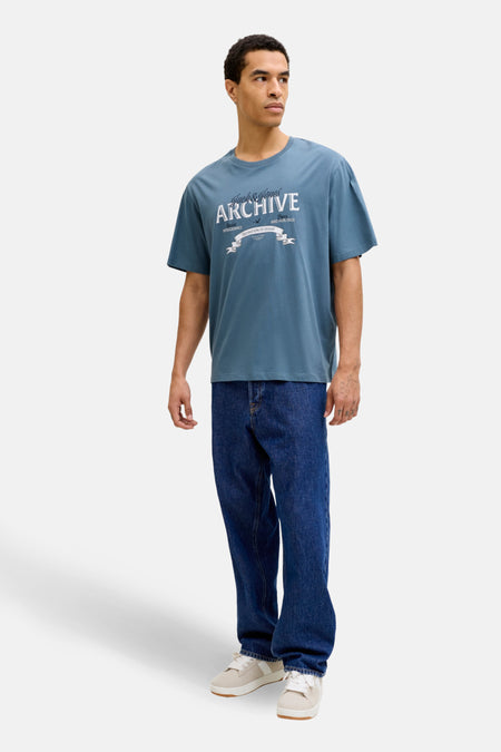 T-shirt bleu à manches courtes de ORIGINALS BY JACK & JONES, avec imprimé texte blanc, porté avec un jean ample et des baskets.