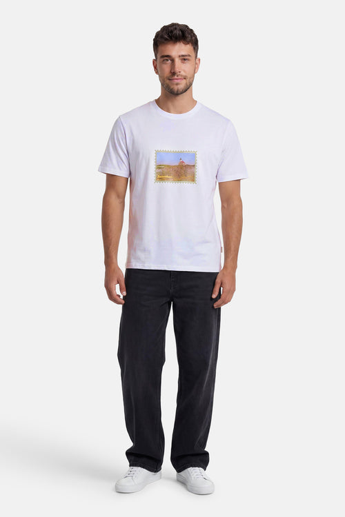 T-shirt blanc de la marque ORIGINALS BY JACK & JONES avec motif timbre paysage et texte, porté avec un jean foncé et des baskets blanches.